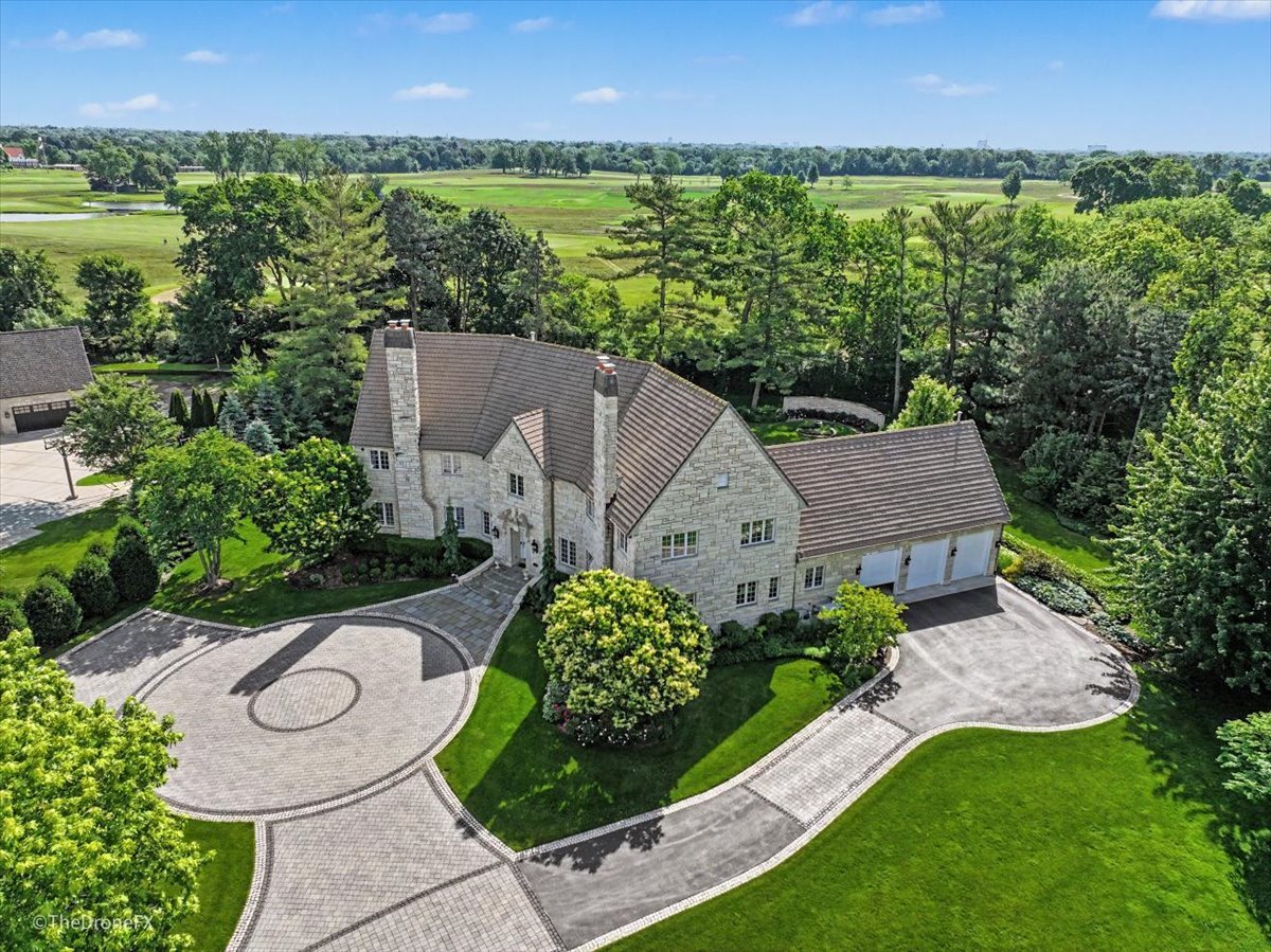 Aerial view of a stone home with a circular driveway and expansive golf-course grounds behind it.