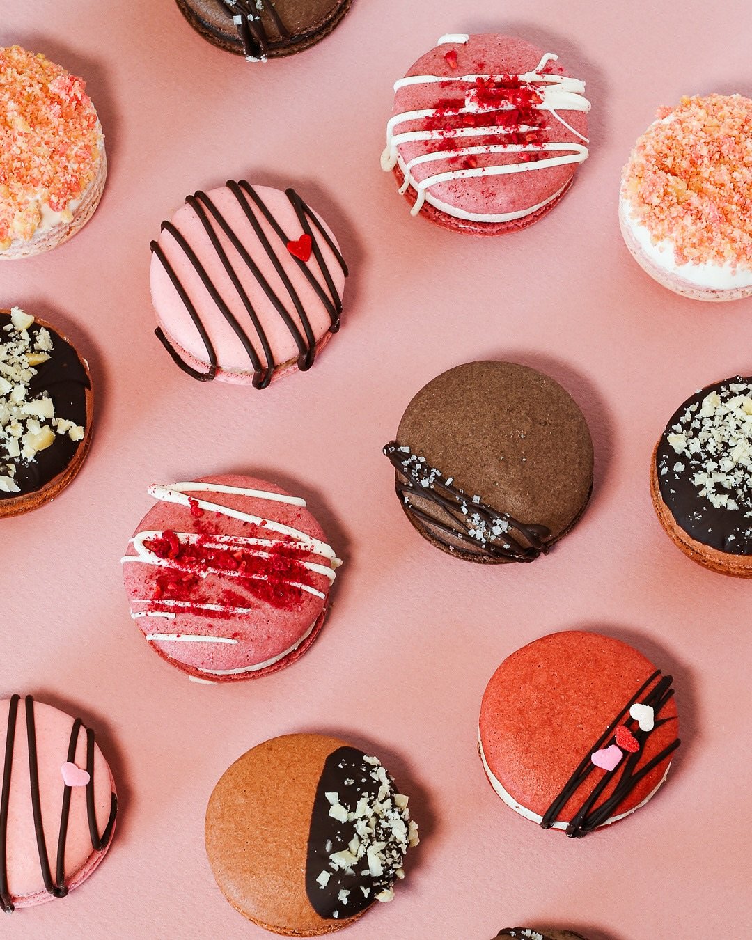 Assorted decorated macarons arranged on a pink surface, with chocolate drizzle, icing, and crumb toppings.