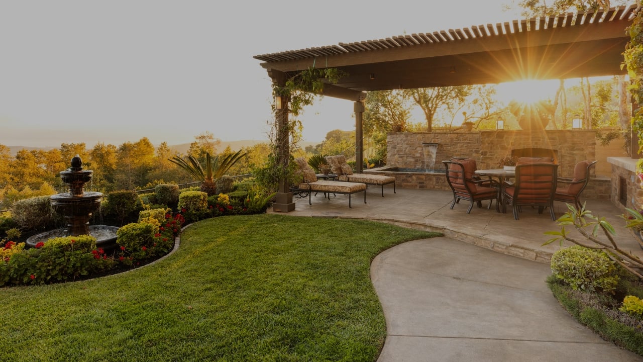 Landscaped backyard patio with a pergola, stone fireplace, fountain, and golden-hour sunlight.
