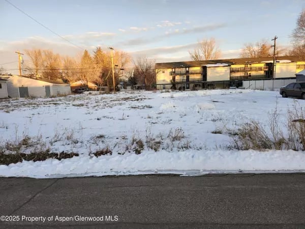 Snow-covered vacant lot beside a road, with low-rise buildings and leafless trees in the background.