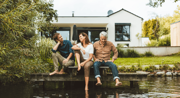 A group of three people sit on a dock with their feet in the water, in front of a modern house surrounded by greenery, conveying a relaxed, serene atmosphere.