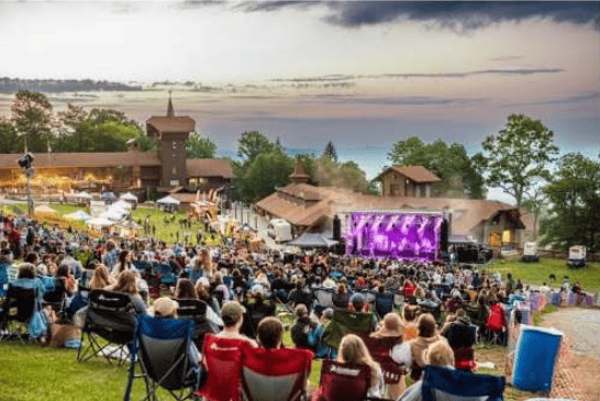 Festival crowd watches a purple-lit stage at a lakeside venue beneath a sunset sky.