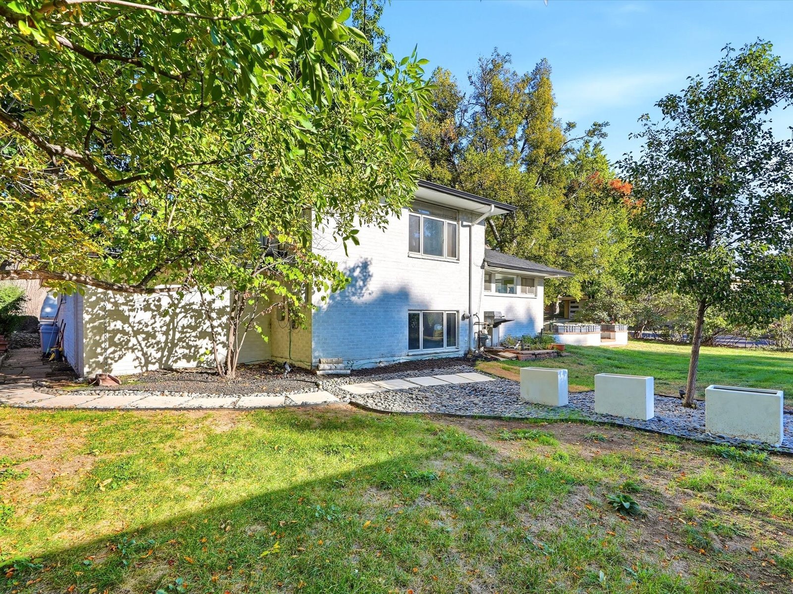 White two-story brick house behind a grassy yard, framed by mature trees and a stone path.