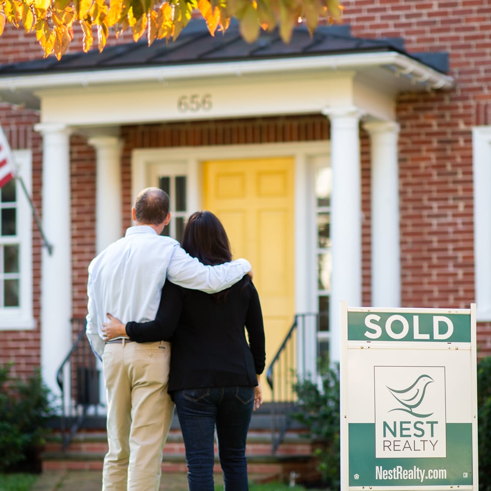 Brick home with a yellow door and SOLD sign as a couple embraces.