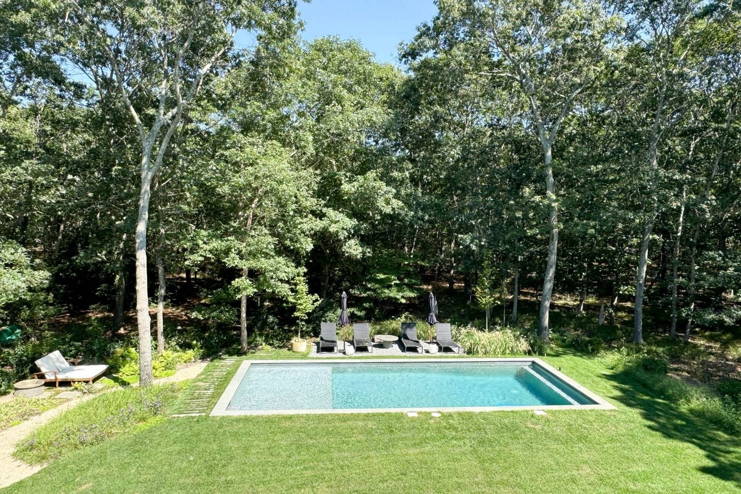 Rectangular backyard pool bordered by lawn and mature trees, with lounge chairs and closed umbrellas.