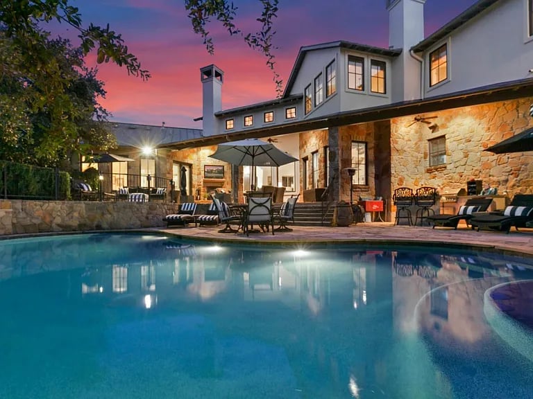 Stone-and-stucco home and furnished patio glowing at dusk beside a swimming pool.