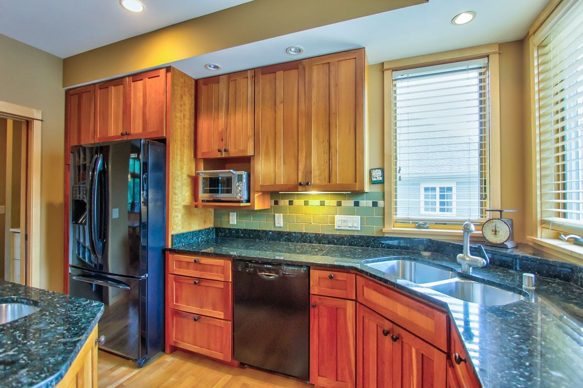 Kitchen with warm wood cabinetry, dark granite counters, green tile backsplash, and a double-basin sink.