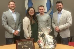 Four adults pose behind a closing gift basket and sign.