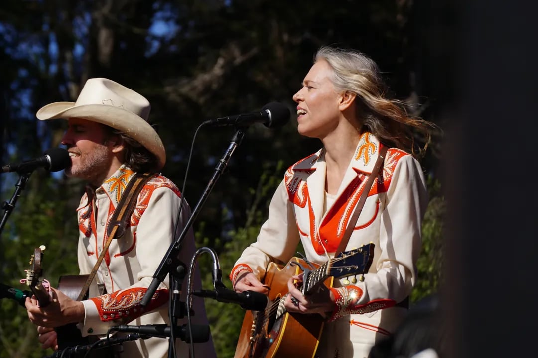 Two musicians performing outdoors in matching cream and orange embroidered western suits, male performer in white cowboy hat playing electric guitar, female performer playing acoustic guitar