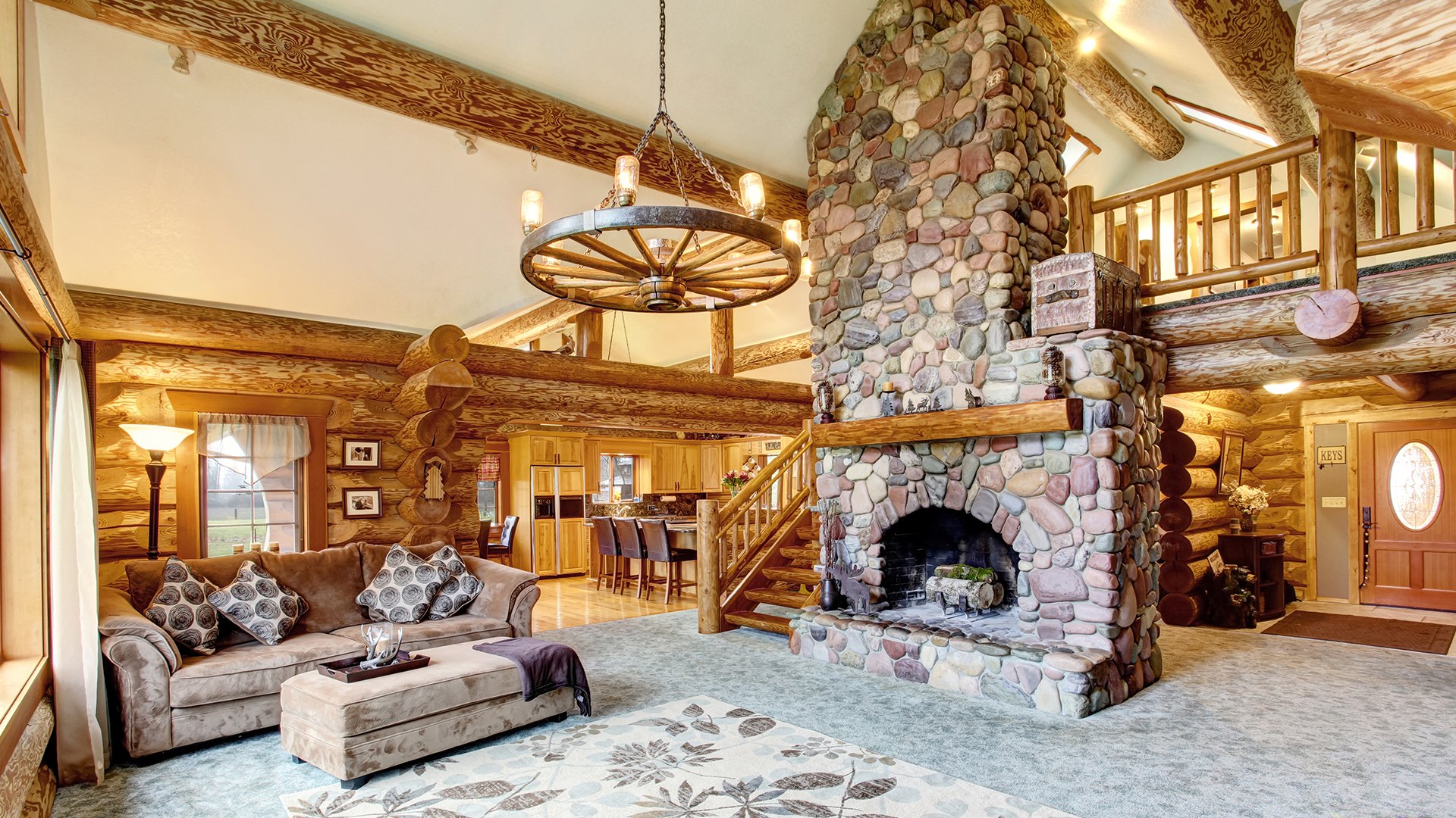 Log-cabin great room with a massive stone fireplace, exposed timber beams, and wagon-wheel chandelier.