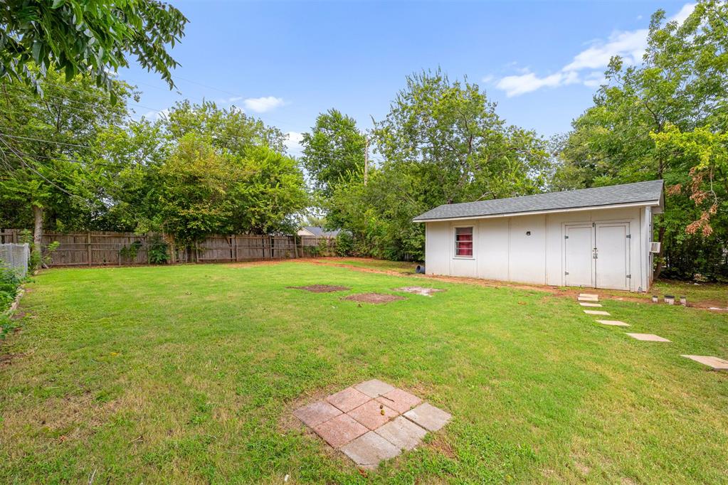 Fenced grassy backyard with a white outbuilding, stepping stones, and mature trees.