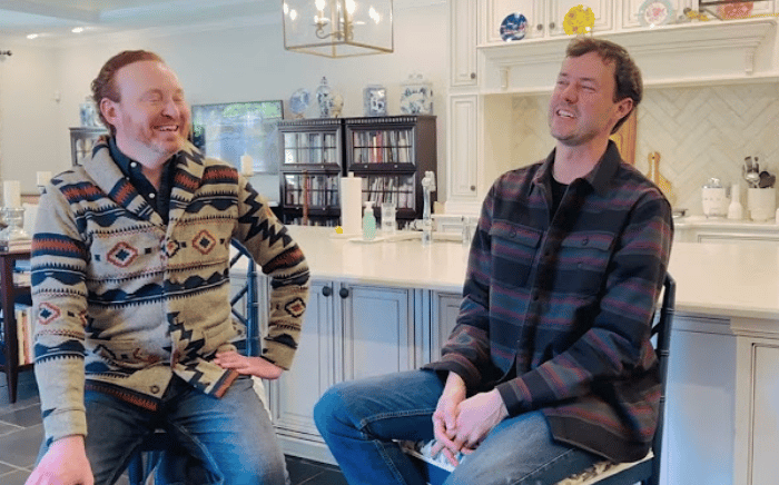 Two men laugh during an interview in a bright kitchen with white cabinetry.