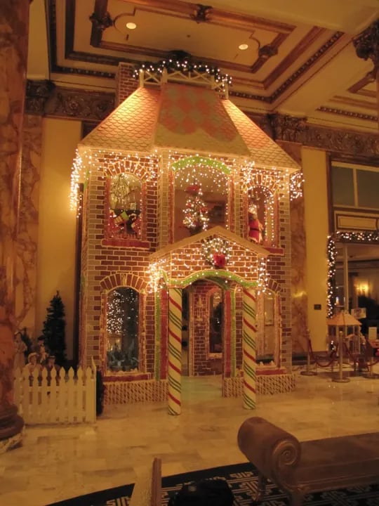 Warmly lit color photo of an enormous, intricately detailed gingerbread house display at the Fairmont Hotel, featuring candy cane columns, twinkling fairy lights, gingerbread elves and Santa inside the windows, a peppermint roof, white picket fence, and surrounding Christmas trees in a grand ornate lobby with marble floors and gilded ceilings.
