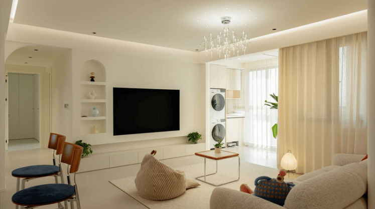 Bright living room with a wall-mounted television, chandelier, and sheer curtains admitting daylight.