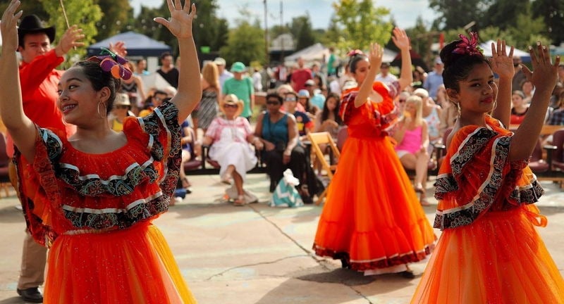 Festival dancers in orange and red dresses perform outdoors before a crowd.