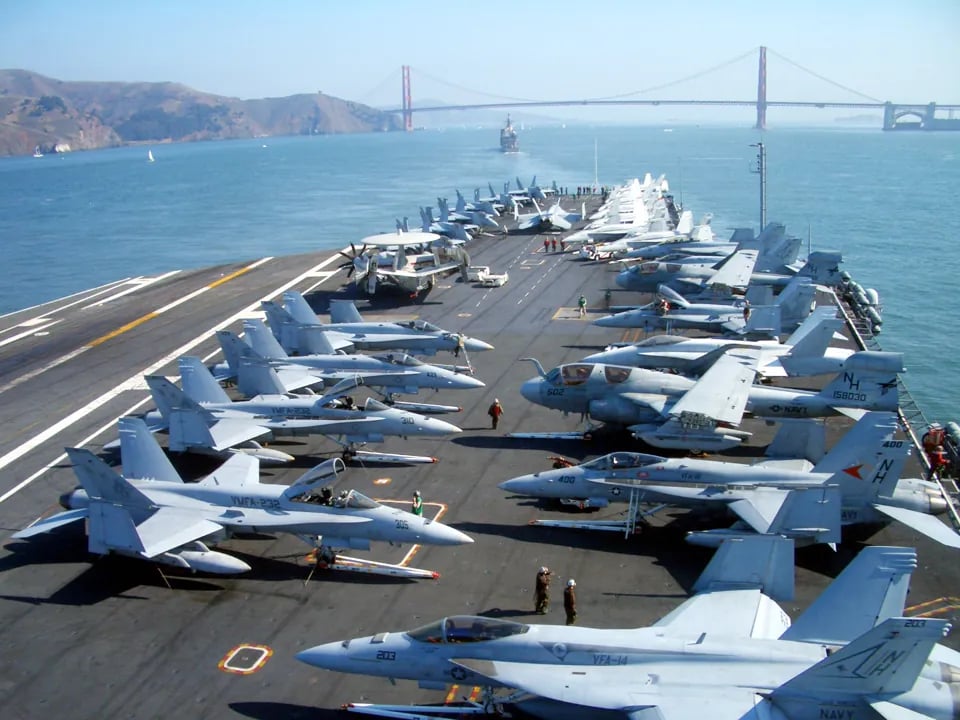 Aircraft carrier flight deck crowded with fighter jets and crew members, Golden Gate Bridge and Marin Headlands visible across San Francisco Bay in background