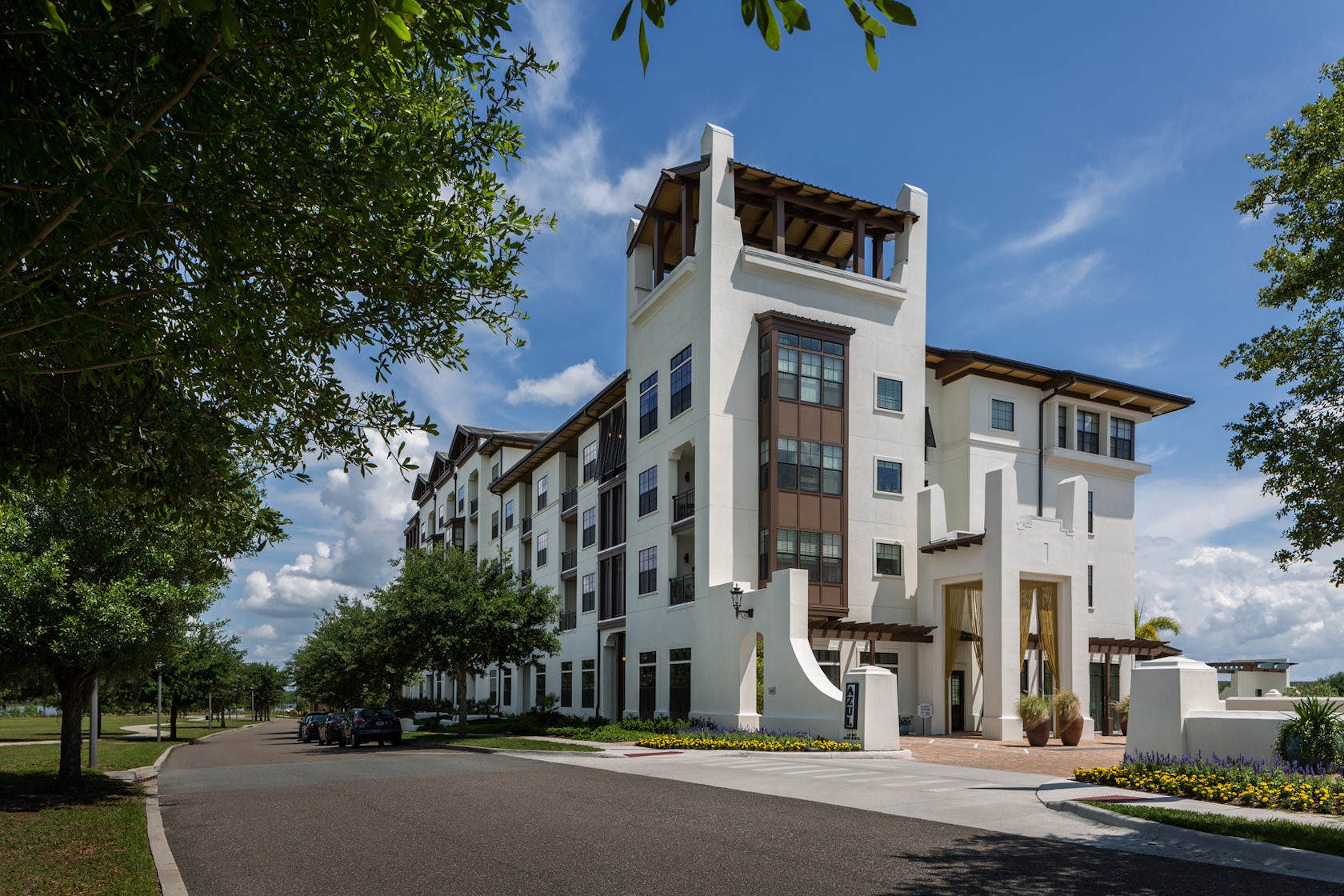 White multi-story apartment building with a rooftop tower, entrance sign, and landscaped flower beds.