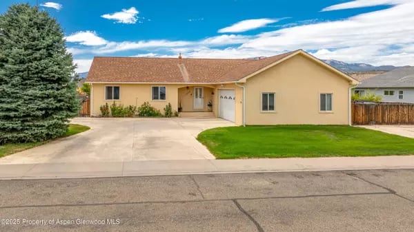 Single-story tan home with an attached garage, wide driveway, green lawn, and large evergreen tree.