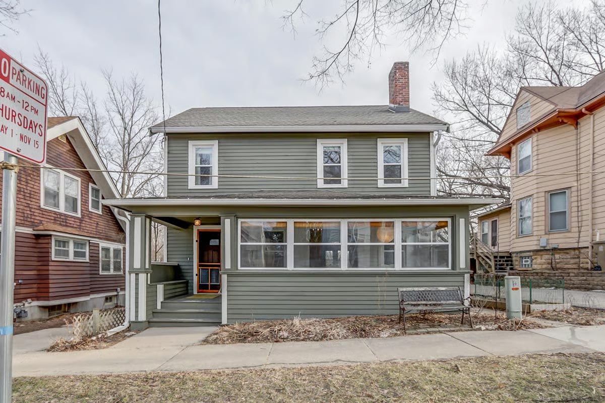 Two-story gray-green house with a wide enclosed porch and brick chimney, between neighboring homes.