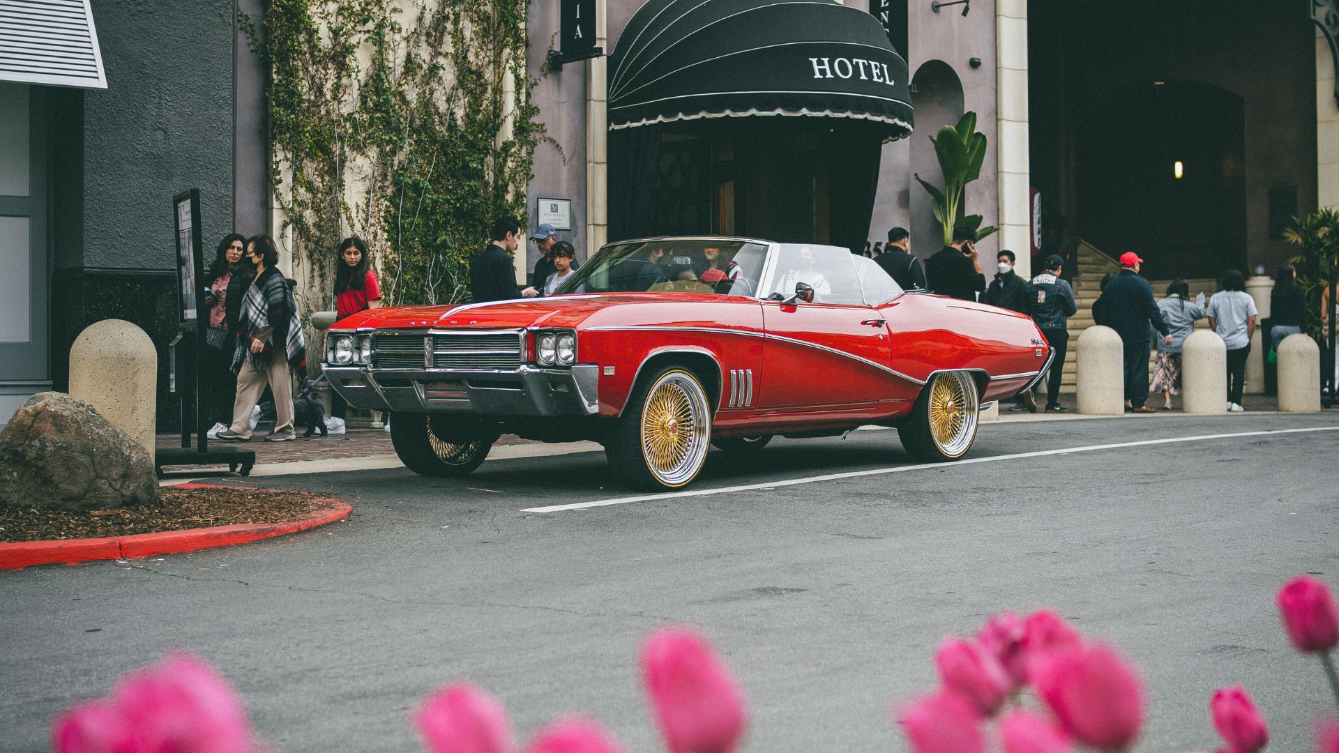 Red classic convertible parked outside a hotel, with pedestrians and pink flowers in the foreground.