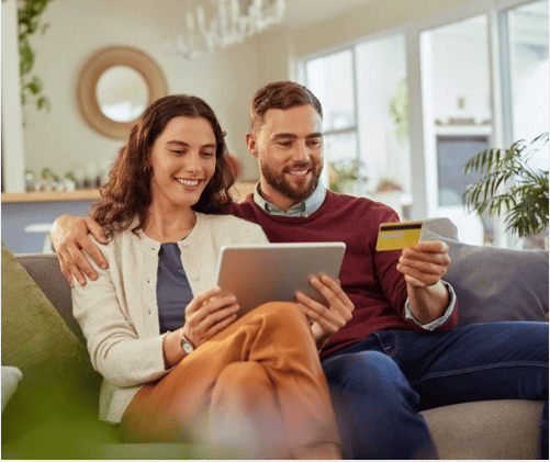 Couple researching a home on a tablet while sitting together on a sofa; one holds a credit card.
