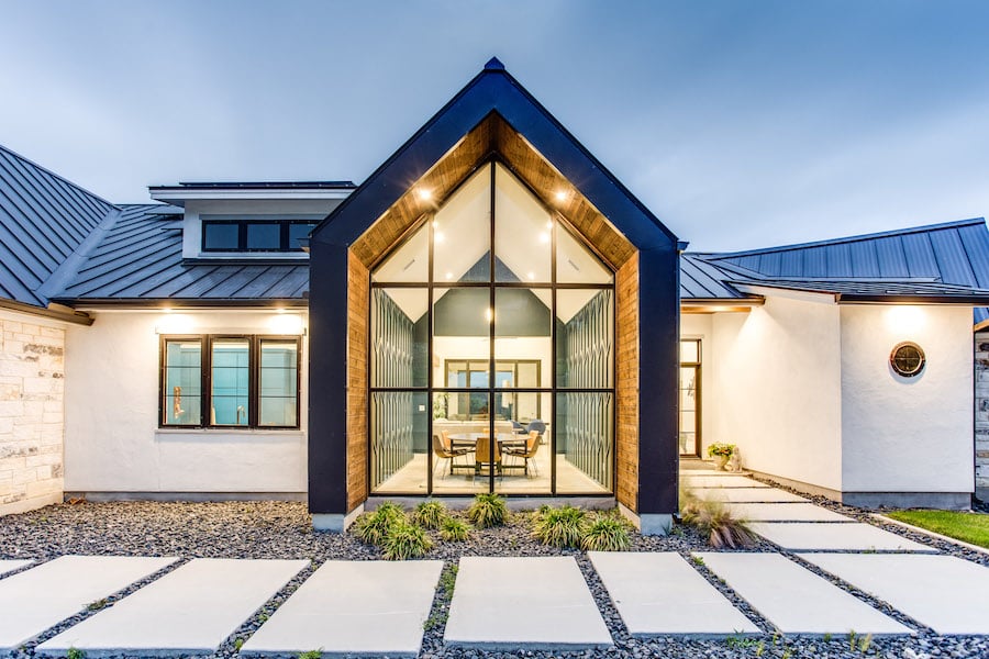 Modern home with a peaked glass entry, metal roof, and broad paved walkway at dusk.
