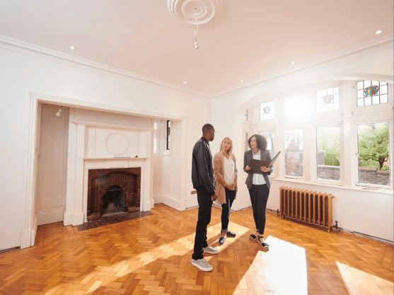Unfurnished living room with a white fireplace, parquet floors, and three people standing by the windows.