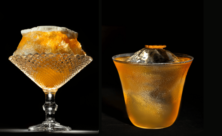 Two amber cocktails, one topped with crushed ice and the other with a large ice sphere.