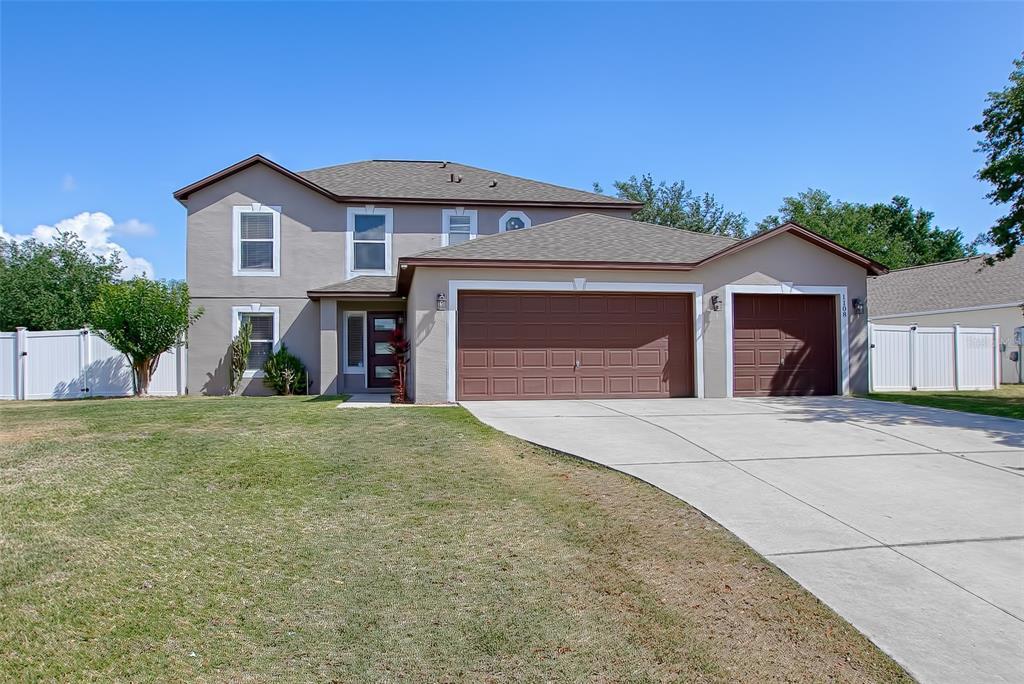 Two-story gray house with brown trim, two garage doors, a wide driveway, and white fencing.