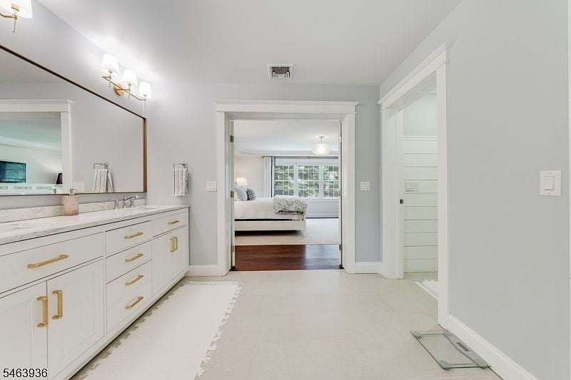 Bathroom with a long white vanity and marble countertop, opening onto a bedroom with broad windows.
