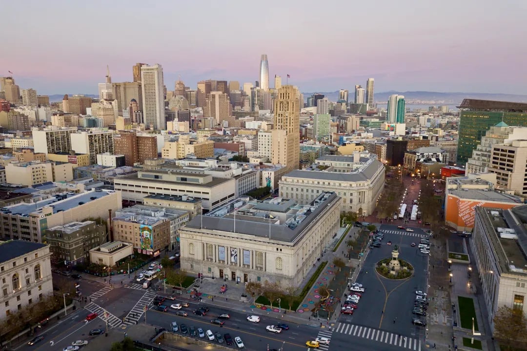 Aerial view of the Asian Art Museum and Civic Center plaza with the downtown San Francisco skyline and Salesforce Tower in the background at dusk.