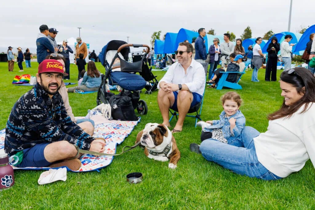 Family with young child and bulldog sitting on grass at outdoor gathering, with blue sculptural art installations and other attendees visible in background at waterfront park