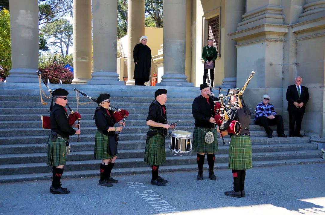 A traditional Scottish pipe band performing at the base of the classical stone columns in San Francisco's Golden Gate Park, drawing an eclectic crowd.