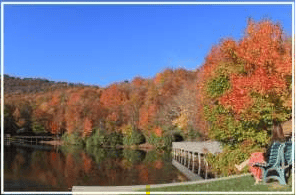 Autumn foliage reflects in a mountain lake beside a dock and colorful Adirondack chairs.