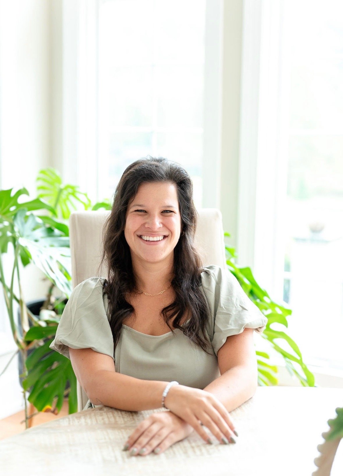 Jordan Robbins smiles seated at a table, with leafy plants and bright windows behind her.