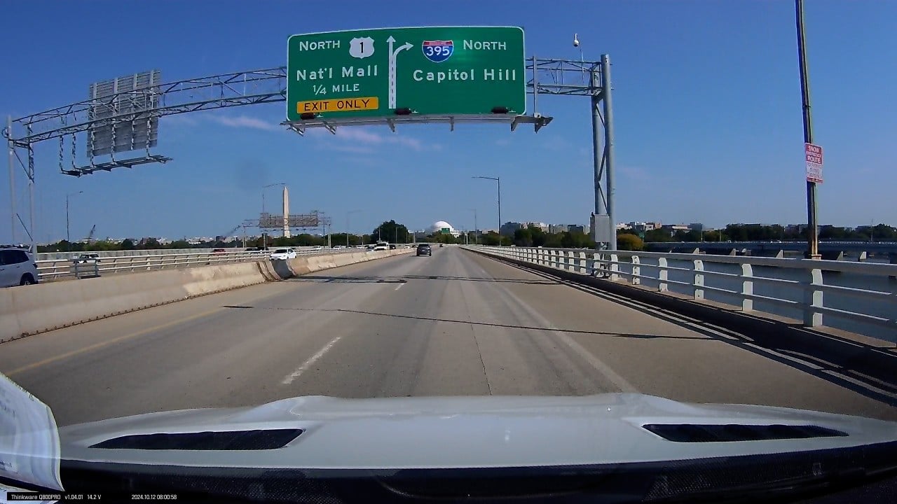 Dash cam view from a vehicle crossing a bridge into Washington, D.C., featuring clear road signs for Capitol Hill and the National Mall. Iconic landmarks such as the Washington Monument and Jefferson Memorial are visible in the background. This image highlights the driver's perspective, emphasizing navigation and the scenic beauty of D.C. traffic routes.