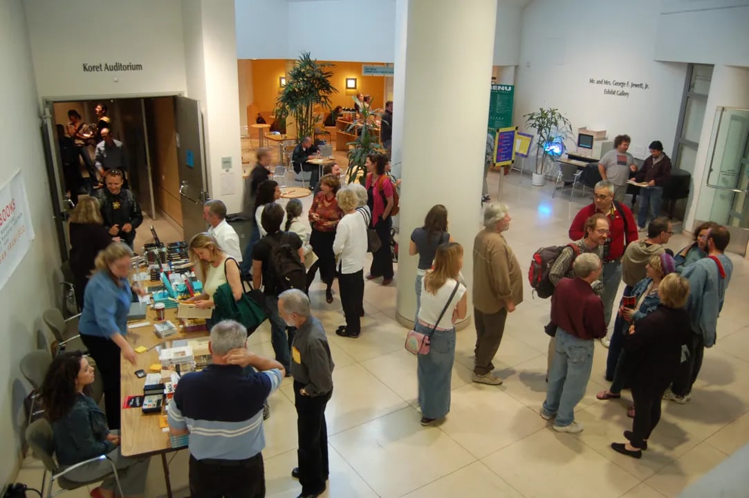 A wide-angle interior view of a brightly lit public space, showing a crowd of people mingling near a book signing or information table on the left, and groups standing around a large central column, with signs for the 