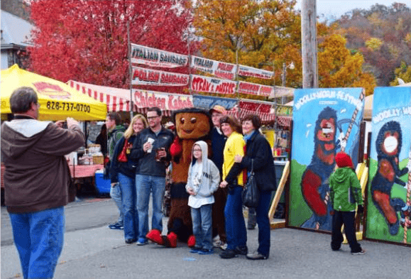 Festival visitors pose with a brown mascot beside food stalls and painted display boards.