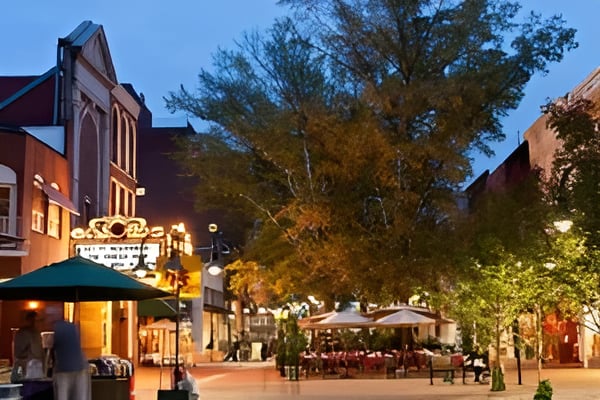 Downtown pedestrian street at dusk with brick storefronts, café seating, and mature trees.