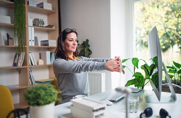 Woman stretching at a home office desk beside a computer and leafy plants.