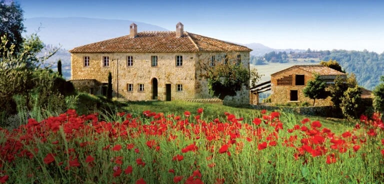 Stone residence with terracotta roof above a field of red poppies, with hills in the distance.