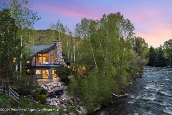 Modern mountain home beside a rushing river, surrounded by trees at dusk.