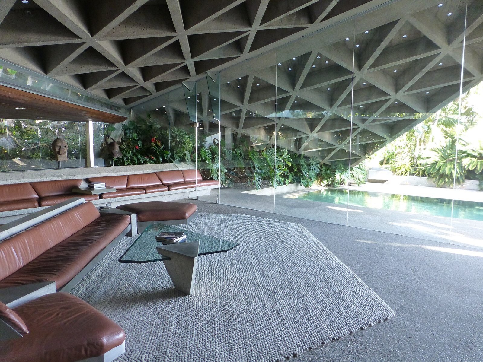 The Sheats-Goldstein living room with a triangular coffered concrete ceiling dotted with small glass skylights, built-in leather seating, and a glass wall opening to the pool.