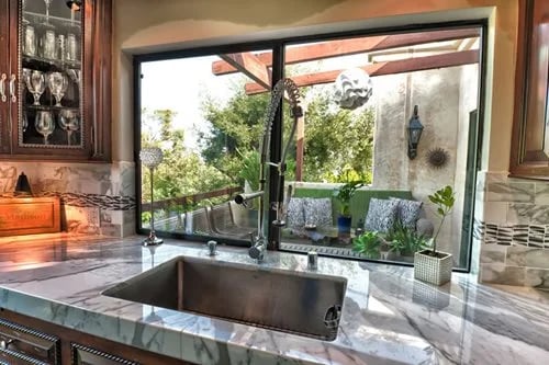Kitchen sink and marble counter beneath a garden-facing window, with a glass-front cabinet and tiled backsplash.