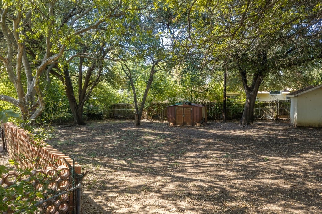 Shaded backyard with mature trees, a small storage shed, and a low brick-and-wire fence.