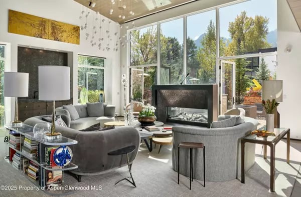 Living room with gray seating, a central fireplace, and floor-to-ceiling windows overlooking trees and mountains.