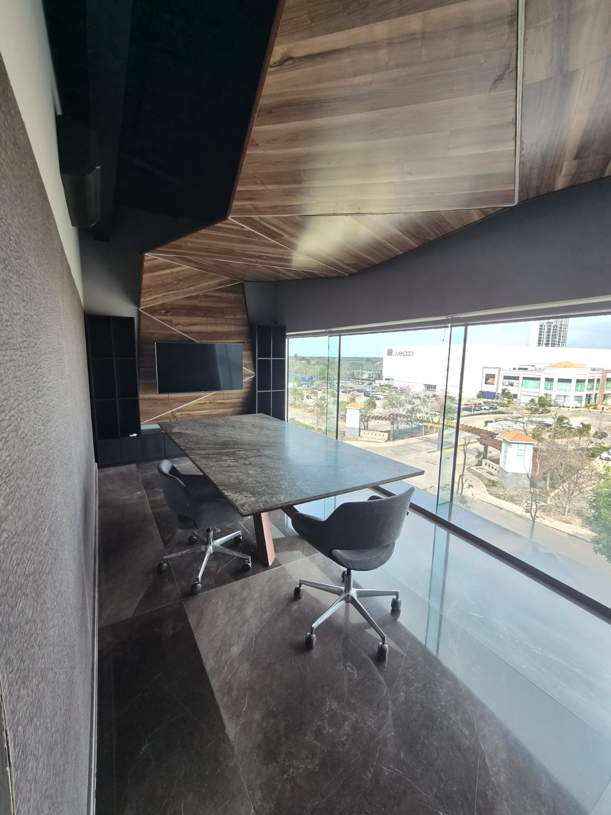 Office meeting room with a conference table and chairs beside floor-to-ceiling windows and geometric wood paneling.