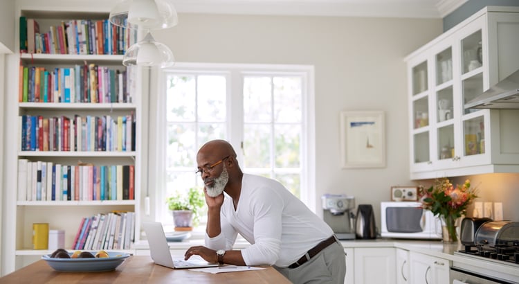 Man reviews a laptop at a kitchen counter beside bookshelves.