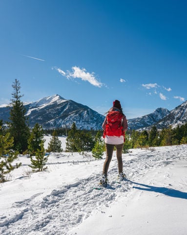 Hiker with a red backpack on a snowy trail facing mountain peaks.