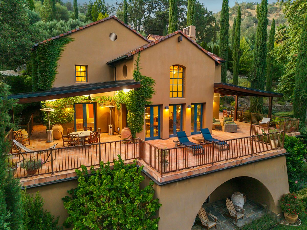 Tan stucco estate with a wide furnished terrace, glowing windows, and cypress trees at dusk.